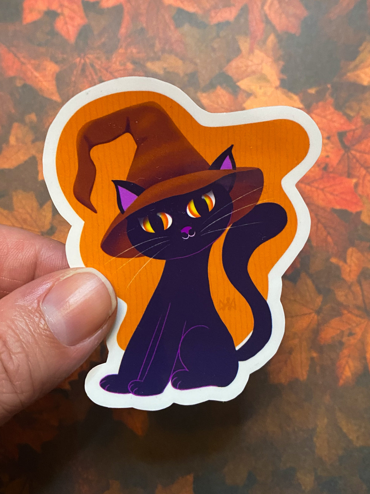 Black cat sticker with orange background held between two fingers to show scale.
